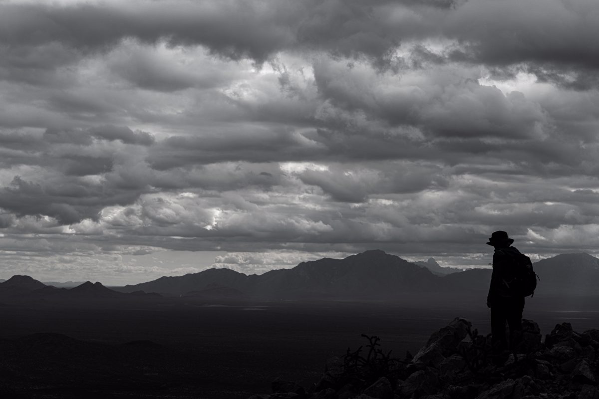 A silhouetted hiker stands on Waterman Peak with clouds over head and distant mountains in the background.