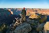 A person stands on shaded rocks in front of a gnarled dead tree with light on the ridges and folds of the opposite side of the Grand Canyon.