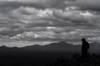 A silhouetted hiker stands on Waterman Peak with clouds over head and distant mountains in the background.