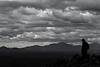 A silhouetted hiker stands on Waterman Peak with clouds over head and distant mountains in the background.