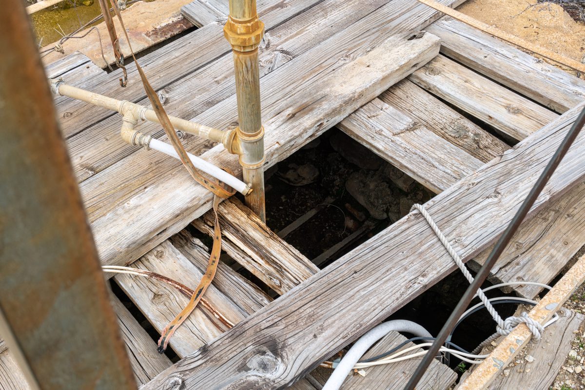 A pipe descends thru a deck of boards partially covering a dark hole below the windmill at Papago Spring.